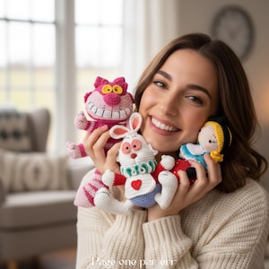 Peut inclure: Trois petites poupées au crochet comprenant un chat rayé rose avec un large sourire, un lapin blanc avec des détails rouges et un cœur, et une poupée aux cheveux jaunes avec une robe bleue. Le texte Page one par err est visible en bas.