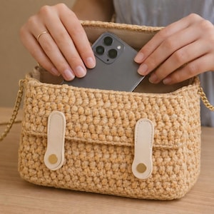 Peut inclure: Un sac à bandoulière en crochet beige avec une chaîne dorée. Le sac a une fermeture à rabat avec deux accents en cuir beige et des boutons dorés. Un smartphone gris est placé à l'intérieur du sac.