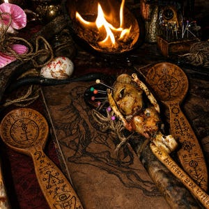 May include: Two wooden spoons with intricate pyrography symbols, a small doll figure pierced with colorful pins, and a rectangular book featuring a dark illustration, all arranged near a burning bowl with visible flames and scattered twine.