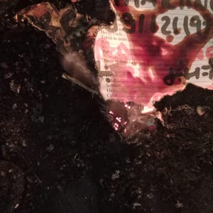 May include: A partially burned document with charred, black edges and a white surface featuring printed text and a large, dark red stain. The visible text includes words like 'Hesap', 'Taklid', 'Riya', 'Tar', 'Yalan', 'İlim', 'Hased', 'Kin', 'Semahet', 'Hicr', and 'Cüret'.