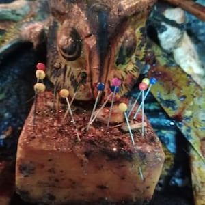 May include: A rectangular, textured orange block serving as a pincushion, topped with a carved wooden owl head featuring dark painted details. Multiple sewing pins with colorful plastic heads in red, yellow, blue, and pink are inserted into the top.