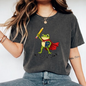 May include: Dark gray t-shirt featuring a cartoon frog superhero holding a pencil and a calculus book. The frog wears a red cape and a blue book with the word "Calculus". The text "WE TEST AT DAWN" is printed below the frog.