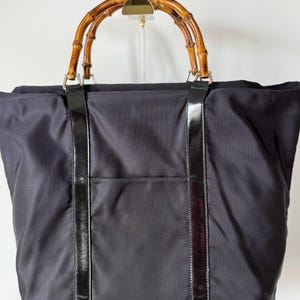 May include: A black nylon tote bag featuring two rounded bamboo handles attached with silver-toned metal hardware. The bag has two vertical black patent leather strips running down the front, which also serve as the attachment points for the handles.