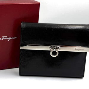 May include: A black leather wallet featuring a polished silver-tone metal bar across the front with a Gancini-style clasp. The wallet is positioned next to a dark red box with the white text Salvatore Ferragamo printed on the front surface.
