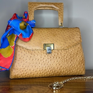 May include: A tan handbag with a textured surface, a rectangular shape, and a handle. A colorful scarf with red, blue, and yellow patterns is draped over the bag. A gold-colored necklace with a circular pendant is in the foreground.