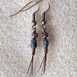 Può includere: Un paio di orecchini fatti a mano con un design unico. Ogni orecchino presenta un gancio color bronzo, un filo a spirale, una piccola perla e dettagli blu scuro e marroni. Gli orecchini hanno un'estetica rustica e bohémien.