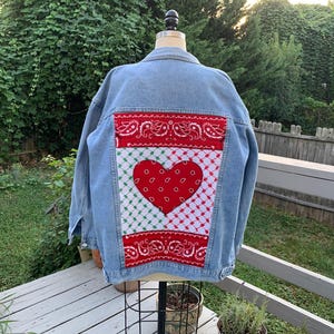 May include: Pale blue denim jacket featuring a red heart and paisley design on the back. The heart is set on a white and green checkered pattern, bordered by red paisley fabric.