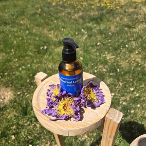 May include: A brown glass bottle of "Ethereal Lotus" body oil with a black pump dispenser sits on a small wooden table. Purple and yellow lotus flowers are arranged around the bottle. The table is outdoors on a grassy surface.