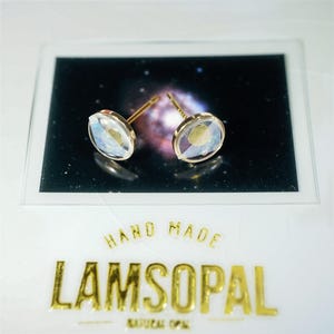 May include: A pair of round stud earrings featuring iridescent, translucent stones set in gold-toned metal. The earrings are displayed on a rectangular card with a space nebula graphic and the gold text HAND MADE LAMSOPAL NATURAL OPAL.
