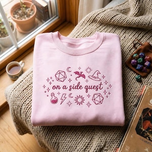 May include: A light pink crewneck sweatshirt featuring dark pink embroidery of the text "on a side quest" surrounded by various icons including a dragon, a wizard hat, a potion bottle, lightning bolts, stars, moons, and two polyhedral dice.