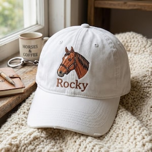 May include: A white baseball cap featuring an embroidered brown horse head wearing a bridle, positioned above the name Rocky in brown text. The cap has a structured crown, a curved brim, and a button at the top, with visible stitching details.