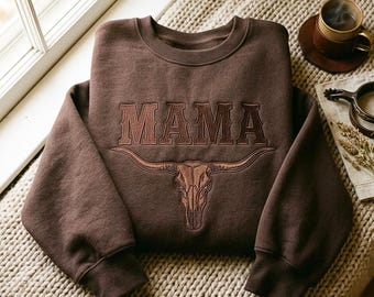 Könnte beinhalten: Ein dunkelbraunes Sweatshirt mit Rundhalsausschnitt, auf dem das Wort MAMA in einer fetten Serifenschrift über einer detaillierten gestickten Illustration eines Longhorn-Stierschädels eingestickt ist. Das Design ist mittig auf der Brust platziert.