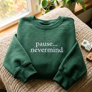 May include: A forest green crewneck sweatshirt featuring the white embroidered text "pause... nevermind" centered on the chest. The garment has long sleeves with ribbed cuffs and a ribbed neckline, showcasing a clean and simple design.
