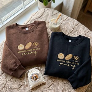May include: A brown and a black crewneck sweatshirt, each featuring an embroidered design of three circular icons depicting bread and wheat, with the text SOURDOUGH RAISING AND JESUS praising. A small white drawstring bag with a logo is included.