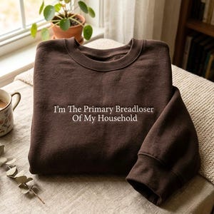 May include: A dark brown crewneck sweatshirt featuring white embroidered text on the chest that reads, I'm The Primary Breadloser Of My Household. The garment has long sleeves, a ribbed neckline, and a solid color finish throughout the fabric.