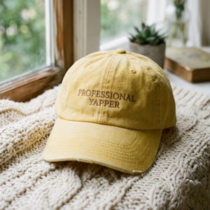 Peut inclure: Une casquette de baseball jaune moutarde avec une finition vieillie et une visière courbée. L'avant de la casquette présente le texte brodé "PROFESSIONAL YAPPER" en fil marron foncé. La casquette a une construction à six panneaux avec un bouton.