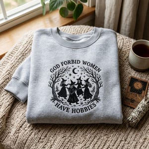 Könnte beinhalten: Ein hellgraues Sweatshirt mit Rundhalsausschnitt und einem schwarzen Grafikdruck von vier Figuren mit spitzen Hüten, die in einem Kreis zwischen kahlen Bäumen, einer Mondsichel und Sternen tanzen, mit dem Text "GOD FORBID WOMEN HAVE HOBBIES".