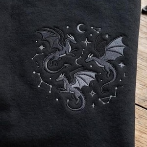 May include: A black fabric garment featuring an embroidered design of three grey dragons in flight. The embroidery includes celestial elements such as a crescent moon, various stars, and constellations arranged in a circular pattern around the dragons.