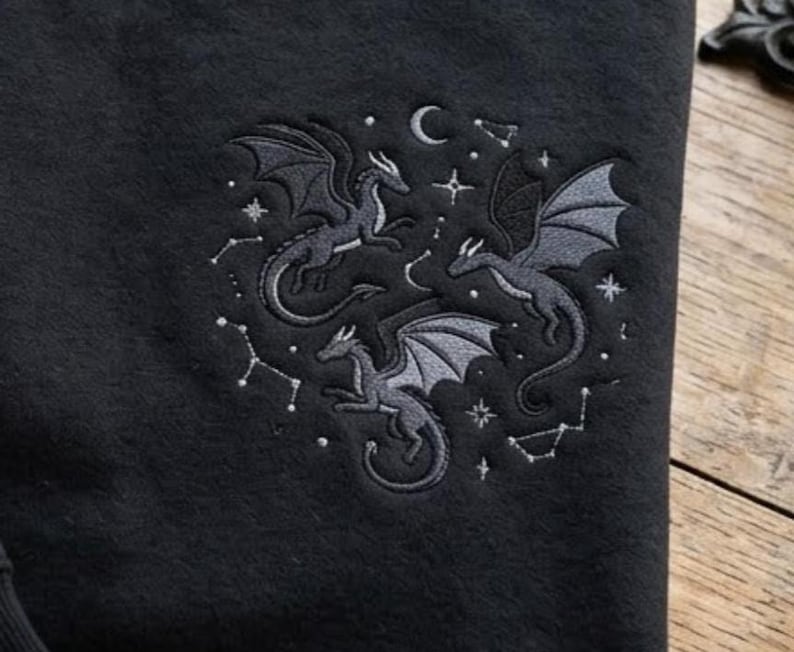 May include: A black fabric garment featuring an embroidered design of three grey dragons in flight. The embroidery includes celestial elements such as a crescent moon, various stars, and constellations arranged in a circular pattern around the dragons.