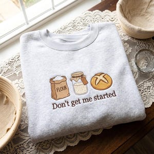 May include: A light gray crewneck sweatshirt featuring an embroidered design of a brown flour bag, a glass jar of sourdough starter, and a round loaf of bread, with the text "Don't get me started" stitched in brown thread below.