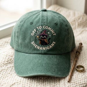May include: A forest green baseball cap featuring an embroidered design of a black cat wearing a purple wizard hat and cape, holding a star-tipped wand. The text "OFF TO COMMIT SHENANIGANS" is embroidered in white around the cat illustration.