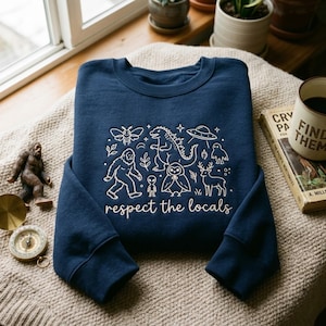 Puede incluir: Una sudadera azul marino de cuello redondo con dibujos de líneas blancas de una polilla, un monstruo, un OVNI, un Bigfoot, un alienígena, un búho y un ciervo. Debajo, el texto "respect the locals" aparece en una fuente cursiva de color blanco.