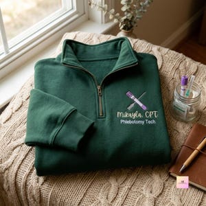 Puede incluir: Una sudadera verde oscuro con cremallera de un cuarto que presenta un bordado de un tubo de ensayo morado y una aguja cruzados en forma de X. Debajo del gráfico, el texto dice Mikayla, CPT y Phlebotomy Tech en letras blancas y amarillo claro.