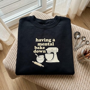 May include: A black crewneck sweatshirt featuring a cream-colored graphic of a stand mixer, a mixing bowl with a whisk, and a rolling pin. The text printed in cream reads, having a mental bake down, positioned above the kitchen appliance illustration.