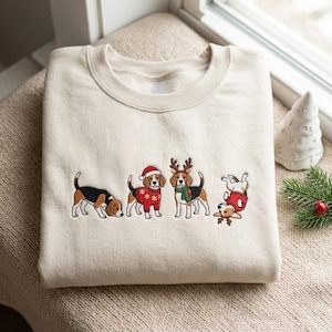 May include: A beige crewneck sweatshirt featuring an embroidered design of four beagle dogs. One beagle sniffs the ground, one wears a Santa hat and red jumper, one wears reindeer antlers and a green scarf, and one rolls on its back wearing antlers.