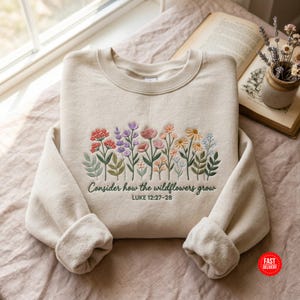 May include: A beige crewneck sweatshirt featuring an embroidered design of colorful wildflowers in shades of pink, purple, orange, and yellow. Below the flowers, the text reads "Consider how the wildflowers grow" and "LUKE 12:27-28".