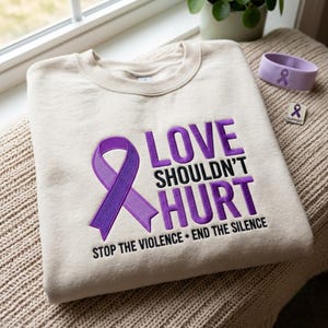 Könnte beinhalten: Ein beigefarbenes Sweatshirt mit Rundhalsausschnitt, einer gestickten violetten Schleife und dem Text "LOVE SHOULDN'T HURT" in Violett und Schwarz sowie dem Zusatz "STOP THE VIOLENCE • END THE SILENCE" darunter.