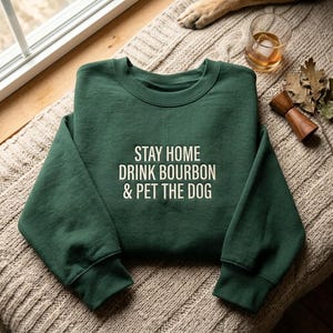 May include: A dark forest green crewneck sweatshirt featuring the white text STAY HOME DRINK BOURBON & PET THE DOG printed in a clean, sans-serif font across the center chest. The garment has long sleeves and a ribbed collar, cuffs, and hem.