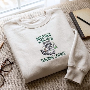 May include: A beige crewneck sweatshirt featuring an embroidered graphic of a raccoon dressed as a scientist holding a green liquid flask. The text above and below the raccoon reads, "ANOTHER WILD DAY TEACHING SCIENCE." The design uses green, gray, and brown thread.