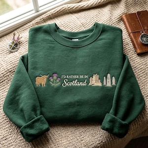 May include: A dark green crewneck sweatshirt featuring embroidered designs of a Highland cow, a purple thistle, a castle, and standing stones. The text "I'D RATHER BE IN Scotland" is embroidered in the center in a mix of block and script fonts.