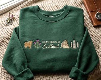 Pode incluir: Uma camisola de gola redonda verde-escura com desenhos bordados de uma vaca das Highlands, um cardo roxo, o texto "I'D RATHER BE IN Scotland", um castelo de pedra e três pedras erguidas dispostas numa linha horizontal no peito.