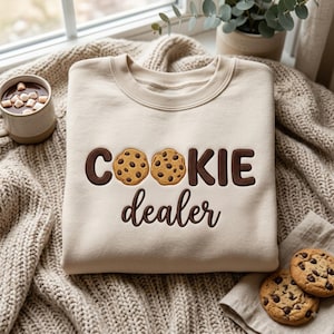 May include: A beige crewneck sweatshirt featuring the embroidered text "COOKIE dealer" in dark brown, with two chocolate chip cookies replacing the letter "O". The design is centered on the chest of the garment, which is made of a soft fabric.