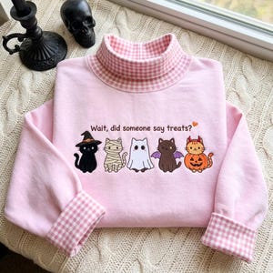 Puede incluir: Una sudadera de manga larga rosa claro con cuello alto con un patrón de cuadros vichy rosa y blanco. El frente muestra un gráfico bordado de cinco gatos con disfraces de Halloween con el texto, Wait, did someone say treats?
