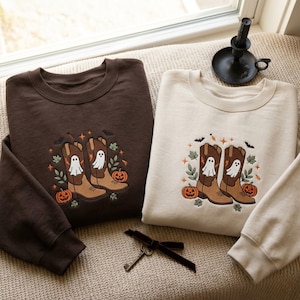 May include: A dark brown sweatshirt and a cream sweatshirt, each featuring an embroidered design of two brown cowboy boots with white ghosts, orange pumpkins, green leaves, small bats, and orange stars centered on the chest area.
