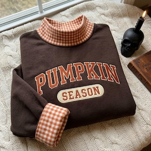 Puede incluir: Una sudadera de color marrón oscuro con cuello alto y puños en un patrón de cuadros vichy naranja y blanco. El frente muestra el texto PUMPKIN SEASON en letras bordadas de color naranja con un borde blanco sobre un fondo color crema.