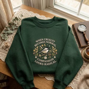 May include: A dark forest green crewneck sweatshirt featuring a central graphic of a small bird perched on a branch, surrounded by floral elements, stars, and a crescent moon, with the text "WHEN CRUELTY BECOMES NORMAL, COMPASSION LOOKS RADICAL."