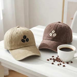 May include: Two baseball caps, one tan with black embroidered coffee beans and one brown with white embroidered coffee beans, positioned next to a white ceramic mug filled with black coffee and scattered roasted coffee beans on a white surface.