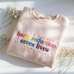 May include: A light beige crewneck sweatshirt featuring the embroidered text "harm reduction saves lives" in a rainbow gradient. The lettering is surrounded by small embroidered stars and hearts in various colors, creating a vibrant design.
