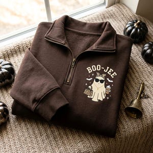 May include: A dark brown quarter-zip sweatshirt featuring a graphic of a ghost wearing sunglasses, a leopard print headband, and a cross necklace. The ghost holds a leopard print handbag and a coffee cup. The text BOO-JEE is printed above.