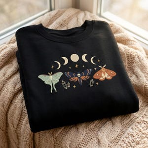 May include: A black crewneck sweatshirt featuring an embroidered design of three moths in shades of light green, dark blue, and brown. The graphic includes a lunar cycle phase arc, small four-pointed stars, and two crystal shapes positioned below.