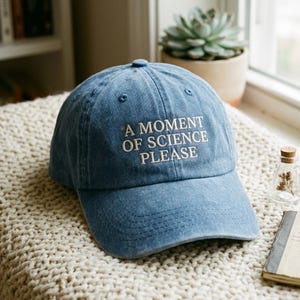 May include: A blue denim baseball cap featuring the embroidered white text A MOMENT OF SCIENCE PLEASE on the front panel. The cap has a structured crown, a curved brim, and a button at the top centre, showcasing a classic six-panel design.