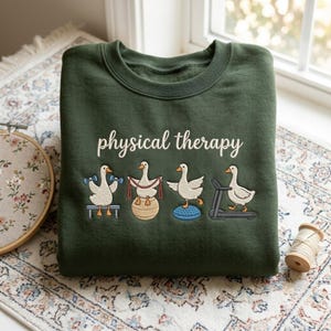 May include: A dark green crewneck sweatshirt featuring the white text physical therapy above an embroidered graphic of four white geese performing various exercises, including lifting dumbbells, using a resistance band, balancing on a ball, and walking on a treadmill.