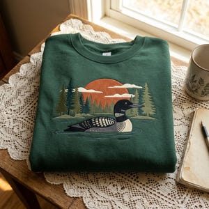 Op de afbeelding: Een donkergroene sweater met ronde hals en een geborduurde afbeelding van een duiker die op een meer zwemt. De scène bevat een grote oranje zon, een dicht dennenbos en witte wolken, allemaal uitgevoerd in gedetailleerd borduurwerk op de stof.