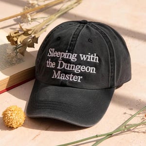 Peut inclure: Une casquette de baseball gris anthracite avec le texte brodé en blanc "Sleeping with the Dungeon Master" sur le panneau avant. La casquette présente une calotte structurée, une visière incurvée et des œillets cousus pour la ventilation.