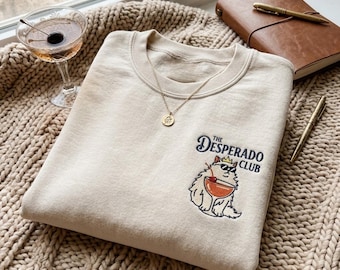 May include: A beige crewneck sweatshirt featuring an embroidered graphic of a fluffy cat wearing sunglasses and a crown, holding a cocktail with a cherry. The text reads THE DESPERADO CLUB. A gold chain necklace with a small pendant rests on the collar.