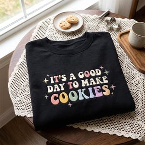 May include: A black crewneck sweatshirt featuring the embroidered text "IT'S A GOOD DAY TO MAKE COOKIES" in a multi-colored, rounded font. The design is accented with small, colorful embroidered star shapes surrounding the lettering.
